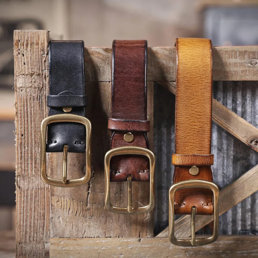 Three leather belts with brass buckles in black, brown, and tan on rustic wood.